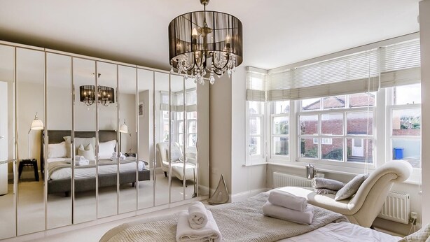 A bedroom with a large bed, a mirror wardrobe, a chandelier, and a view of the outside through large windows.