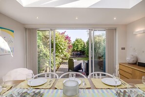A dining area with a table set for four, a sliding glass door leading to a garden, and a white cabinet with a vase. - 4 Bed in Southwold (oc-aalavh) (Southwold)