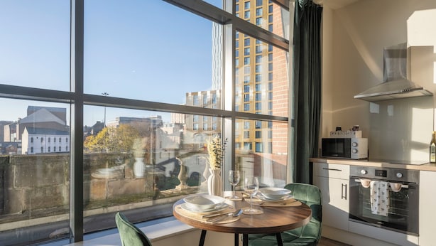 Une cuisine moderne dotée d’une grande fenêtre, un coin repas avec des chaises vertes et une vue sur le paysage urbain.