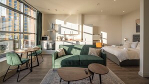 Une chambre d’hôtel moderne avec un canapé vert, un lit, un coin repas avec une table et des chaises, et une cuisine en arrière-plan.