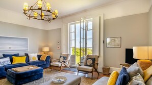A spacious living room with a blue sofa set, a grey ottoman, and a wooden chair. A chandelier hangs from the ceiling, and there is a large window providing a view of the outdoors. - 5 Bed in Southwold (oc-aa3cen) (Southwold)