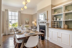 Une salle à manger avec une longue table en bois dressée pour six personnes, une cheminée et une bibliothèque agrémentée d’objets décoratifs.