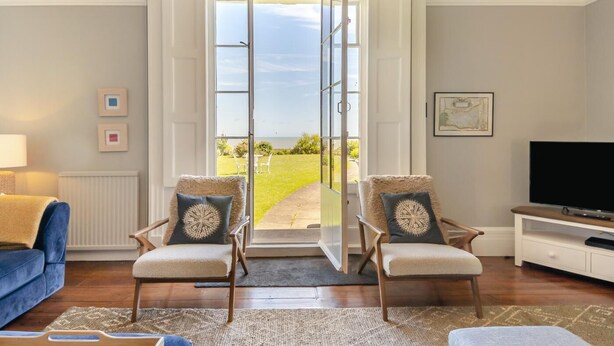 A living room with a large sliding glass door leading to a patio. There are two chairs with decorative pillows, a sofa, a TV on a stand, and a lamp. - 5 Bed in Southwold (oc-aa3cen) (Southwold)