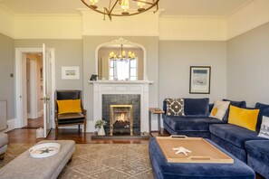A living room with a fireplace, a blue sofa set, a patterned rug, and a wooden coffee table. - 5 Bed in Southwold (oc-aa3cen) (Southwold)