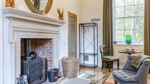 A living room with a fireplace, a green chair, a table, and a window with a view of trees. - 1 Bed in Wells-next-the-Sea (oc-1939) (Wells-next-the-Sea)