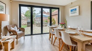 A dining area with a large table set for six, a wicker chair, and a view of the backyard through glass doors. - 4 Bed in Brancaster (oc-1918) (Brancaster)