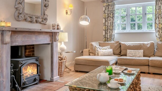 A cozy living room with a fireplace, a sofa set, a glass coffee table, and a large window with curtains. - 4 Bed in Brancaster (oc-1918) (Brancaster)