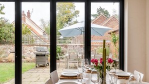 An outdoor dining area with a table set for six, a grill, and a view of a brick house and garden. - 4 Bed in Brancaster (oc-1918) (Brancaster)
