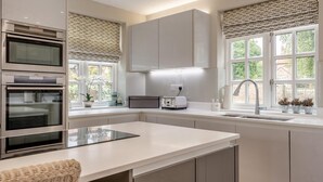 A modern kitchen with built-in ovens, a central island, and a window with a view of greenery. - 4 Bed in Brancaster (oc-1918) (Brancaster)