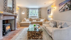 A cozy living room with a fireplace, a sofa set, a glass coffee table, and a painting on the wall. - 4 Bed in Brancaster (oc-1918) (Brancaster)