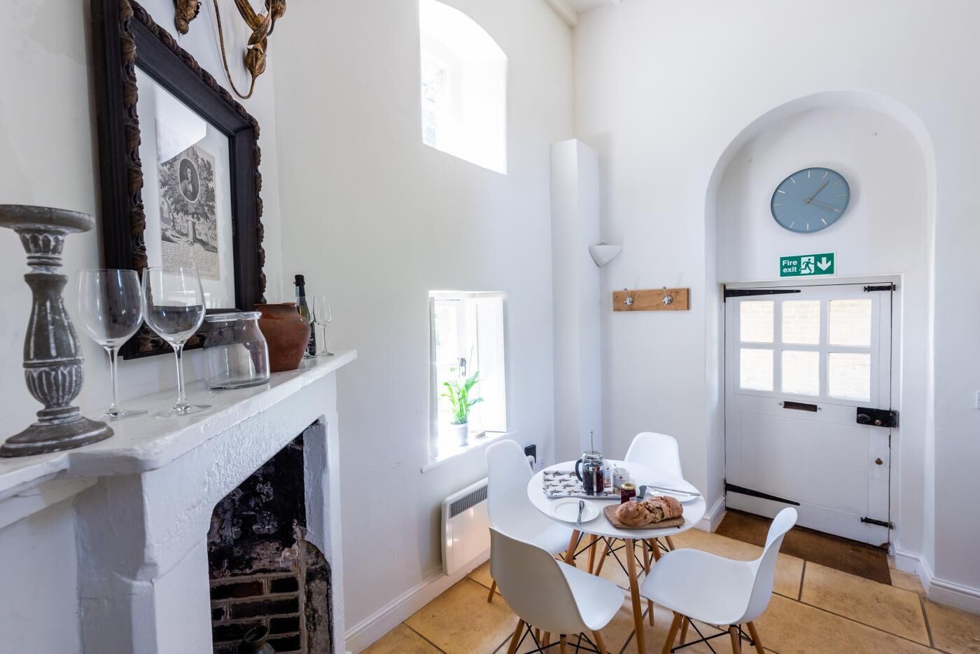 Un coin repas avec une table blanche, des chaises et une cheminée. Il y a une horloge au mur et une porte portant un panneau indiquant la sortie de secours en cas d’incendie.