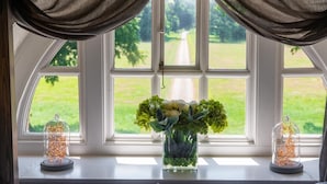 Ein Fensterblick auf eine grüne Landschaft, eine Vase mit Blumen und zwei Glasornamente auf dem Fensterbrett.