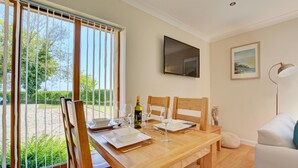 A dining area with a wooden table set for two, a bottle of wine, and a television mounted on the wall. - 2 Bed in Ingworth (oc-1731) (Ingworth)