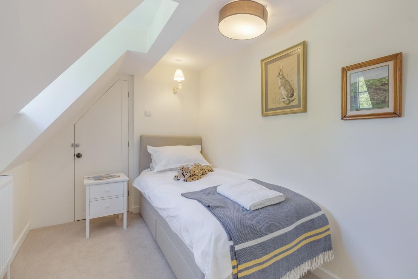 A bedroom with a skylight, a bed with a blanket, a nightstand, and two framed pictures on the wall.