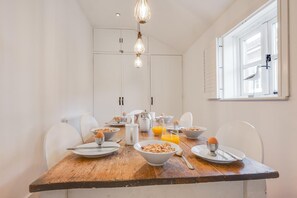 A dining area with a wooden table set for breakfast, white chairs, and a window with blinds. - 3 Bed in Wells-next-the-Sea (oc-1963) (Wells-next-the-Sea)