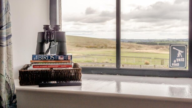 A window with a view of a rural landscape, a telescope, and a stack of books on a windowsill. - 1 Bed in Powburn (oc-nb662) (Ilderton)