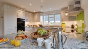 A modern kitchen with white cabinets, a marble countertop, and a breakfast setup including fruits, croissants, and coffee. - 3 Bed in St. Merryn (oc-p00725) (Saint Merryn)