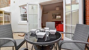 A round glass table with wicker chairs, a red sofa, and a white door leading to a room with a red brick wall. - 4 Bed in Cromer (oc-1942) (Cromer)