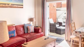 A cozy living room with a red leather sofa, a wooden coffee table, and a view of the patio area with wicker furniture and a table. - 4 Bed in Cromer (oc-1942) (Cromer)