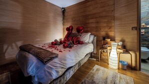 Ein Schlafzimmer mit einer Wand aus Holzlatten, einem Bett mit weißer Bettwäsche, roten, herzförmigen Ballons, die 'LIEBE' buchstabieren, und einem kleinen Nachttisch mit einer Lampe.
