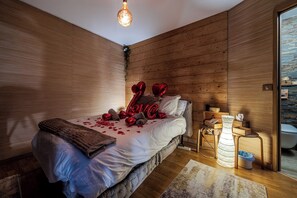 Een slaapkamer met een wand van houten latten, een bed met wit beddengoed, rode hartvormige ballonnen waarop 'LOVE' staat, en een klein houten nachtkastje met een lamp.
