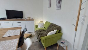 Un espace de vie compact comprenant un canapé vert, une petite table, un téléviseur posé sur un meuble en bois et un meuble blanc.
