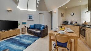 Een moderne woonkamer met een flatscreen televisie, een bank, een eettafel met stoelen, een keuken met apparatuur en een groot raam dat voor natuurlijk licht zorgt.