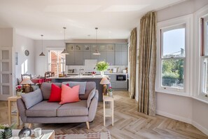 Uma sala de estar moderna com um sofá cinzento, uma área de jantar com toalha de mesa vermelha e uma cozinha com armários cinzentos e iluminação pendente.