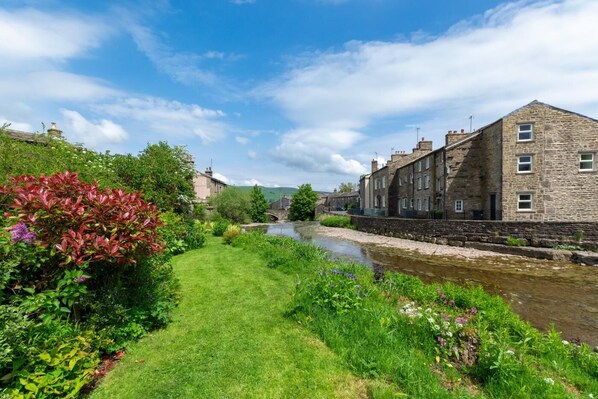 A riverside view with stone buildings, a grassy path, and lush greenery.