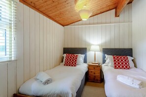 A room with two beds, wooden headboards, a bedside table with a lamp, and a window with blinds.