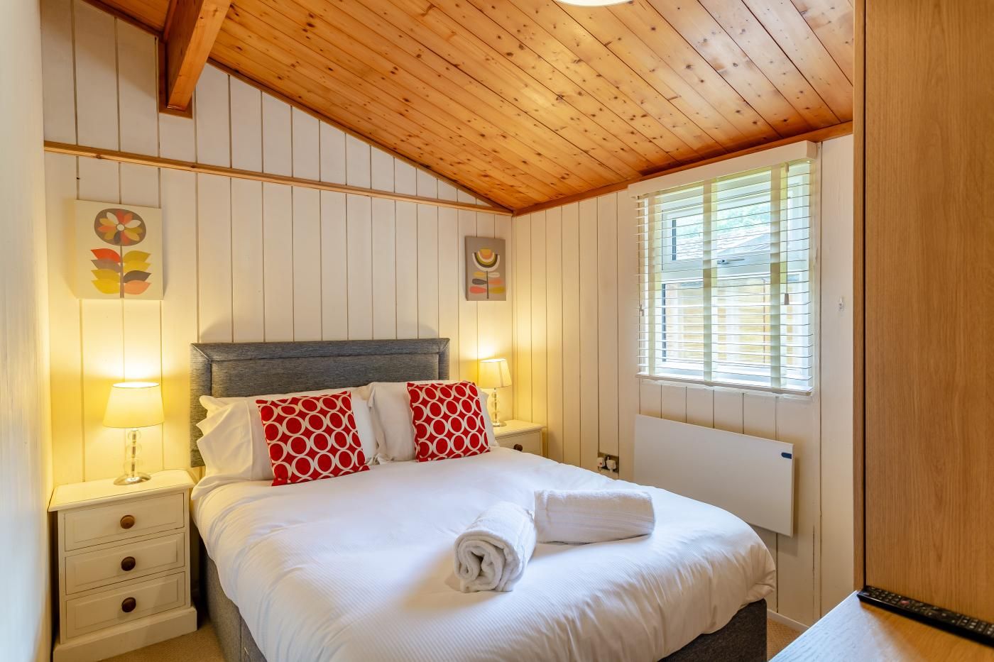 Une chambre avec un plafond en bois, un lit avec une literie blanche et des oreillers rouges, une table de chevet avec une lampe, et une fenêtre avec des stores.