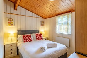 A bedroom with a wooden ceiling, a bed with white bedding and red pillows, a bedside table with a lamp, and a window with blinds.