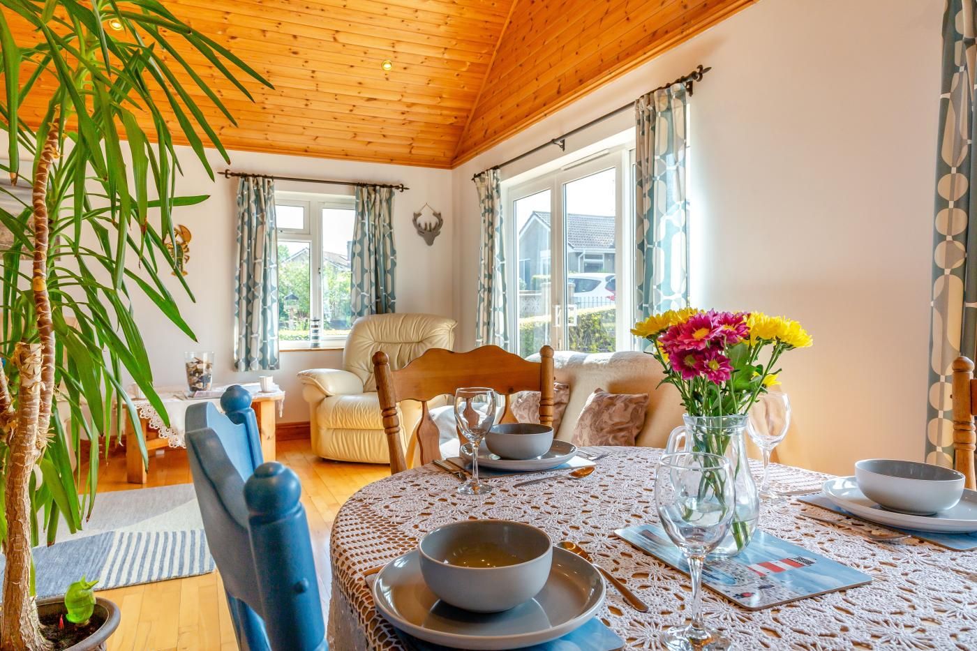 Een gezellig eetgedeelte met een ronde tafel gedekt voor twee personen, een vaas met bloemen en een uitzicht op de omgeving door grote ramen.
