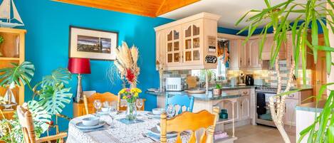 A dining area with a table set for four, surrounded by wooden chairs, a blue wall, and a kitchen in the background.