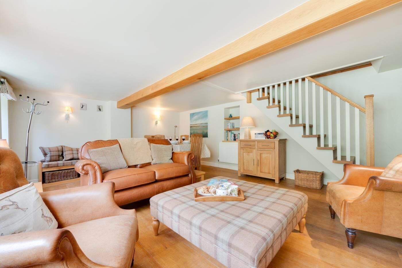 A cozy living room with a brown leather sofa, a plaid ottoman, and a wooden staircase.