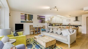 A modern living room with a sofa, coffee table, and TV. There is a kitchen area with cabinets and a dining table. - 4 Bed in Southwold (oc-hswall) (Southwold)
