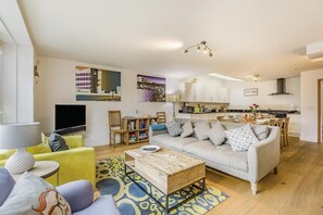 A modern living room with a sofa, coffee table, and TV. There is a kitchen area with cabinets and a dining table.