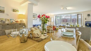 Comedor con mesa de madera dispuesta para una comida, un florero con flores y vistas al exterior a través de amplias ventanas.