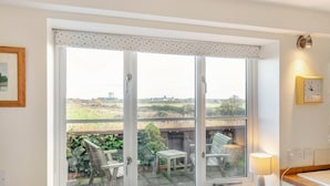 A room with a large window, a table with chairs, a lamp, and a clock on the wall. - 3 Bed in Southwold (oc-hshore) (Southwold)