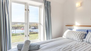 A hotel room with a bed, curtains, a view of boats on water, and a headboard. - 3 Bed in Southwold (oc-hshore) (Southwold)