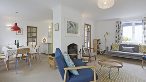 A living room with a sofa, a dining table set for two, a fireplace, and a large window with curtains. - 5 Bed in Walberswick (oc-hferin) (Walberswick)