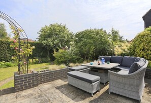 Eine Terrasse im Freien mit Korbmöbeln, einer Ziegelwand und einem Garten mit verschiedenen Pflanzen und Blumen.