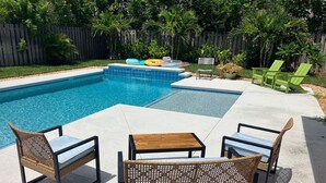 Un patio trasero con piscina, dos sillas de mimbre, una mesa de madera y una pequeña zona de terraza.