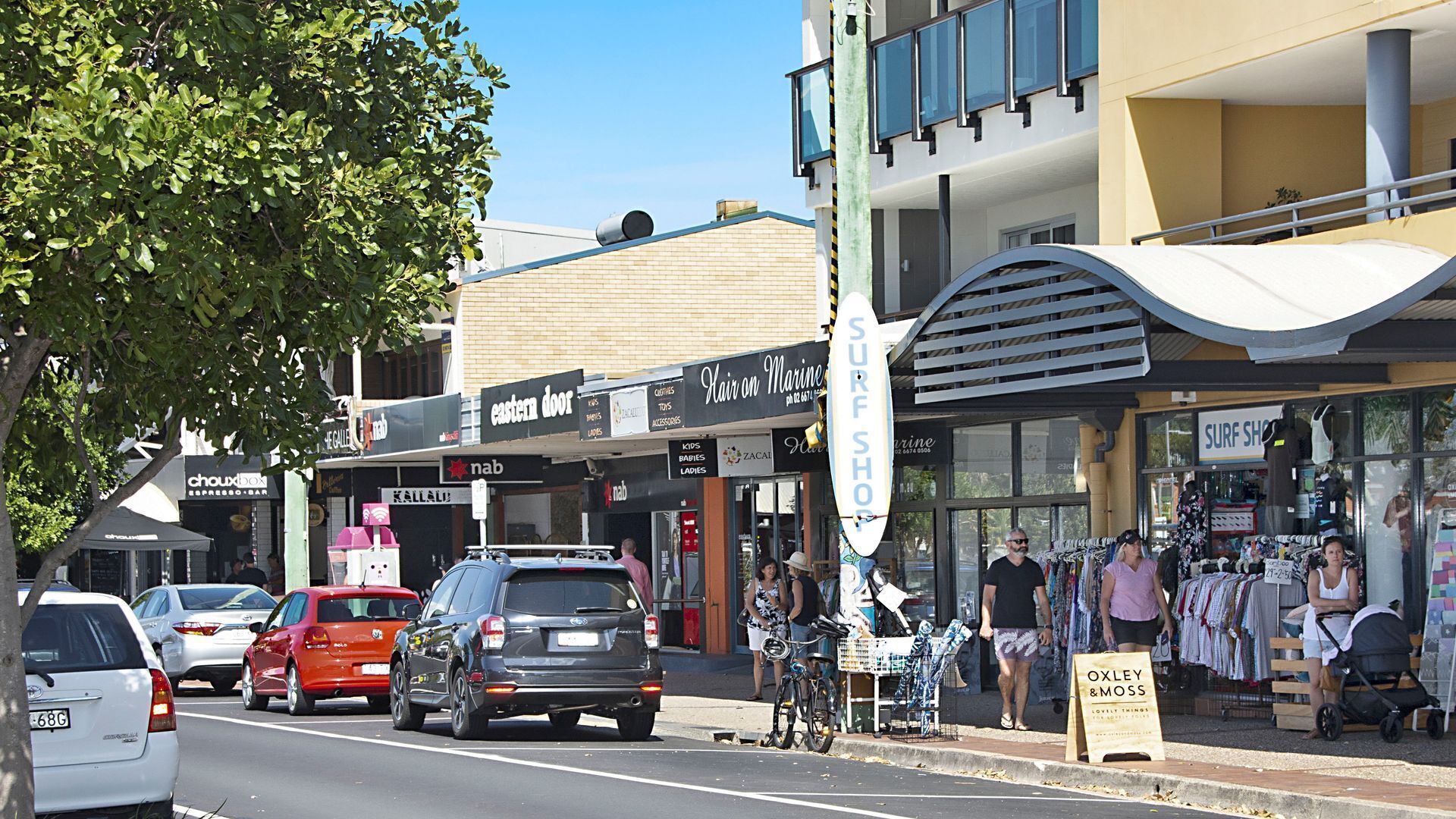 A street view with shops, including 'Hair on Marine' and 'Surf Shop'. Cars are parked along the street, and there are pedestrians.