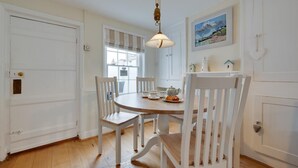 Een eetruimte met een ronde tafel, witte stoelen, een hanglamp en een raam met uitzicht op zee.