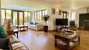 Ein geräumiges Wohnzimmer mit Sofa, Couchtisch und Fernseher. Große Fenster und ein Kamin sind ebenfalls vorhanden.
