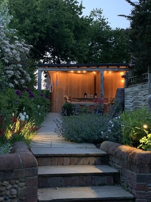 Una pérgola de madera con luces colgantes, un muro de piedra y un jardín con diversas plantas y flores.