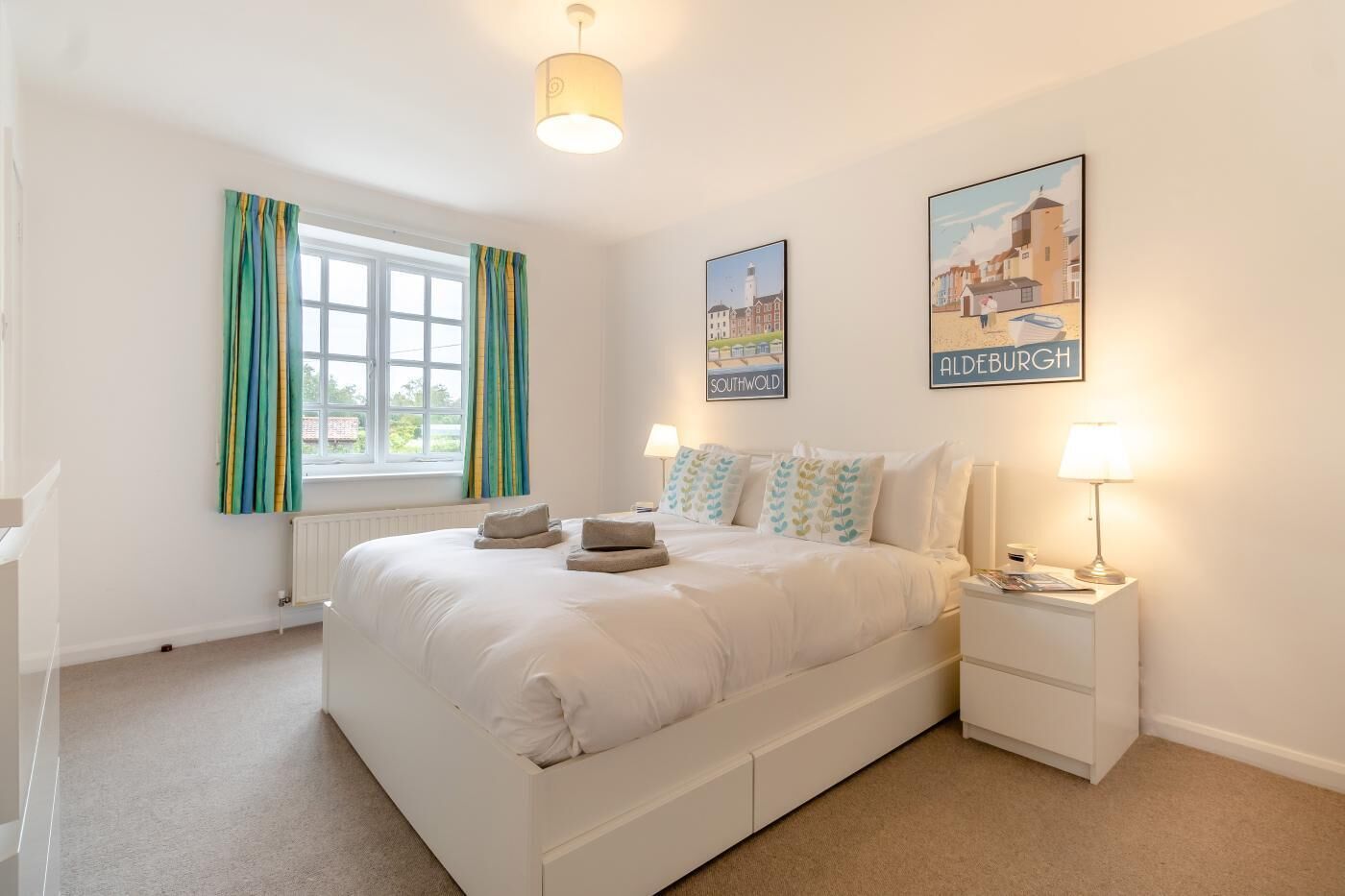 A bedroom with a bed, two bedside tables, a window with curtains, and two wall-mounted pictures.
