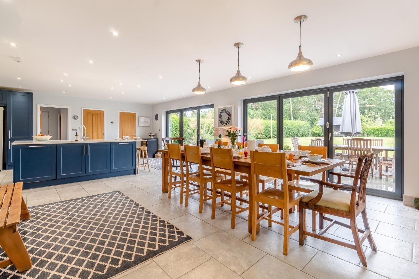 A modern kitchen with blue cabinets, a dining area with wooden chairs, and large windows offering a view of the outdoors.