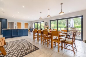 A modern kitchen with blue cabinets, a dining area with wooden chairs, and large windows offering a view of the outdoors. - 5 Bed in Walberswick (oc-hlodg2) (Walberswick)